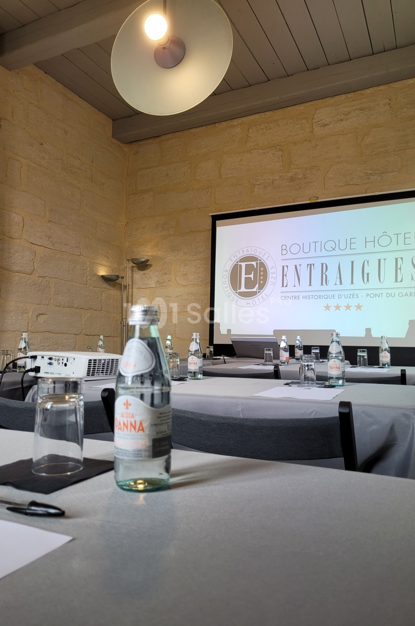 Salle de réunion avec tables dressées, bouteilles d'eau, verres, projecteur et écran affichant un logo d'hôtel.