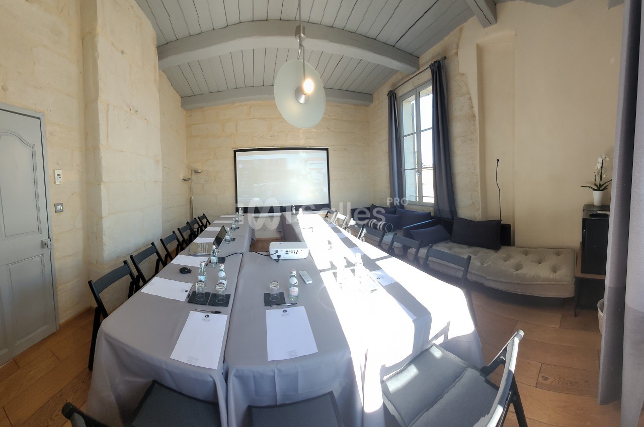 Salle de réunion lumineuse avec une grande table rectangulaire, chaises noires, écran de projection et murs en pierre claire.