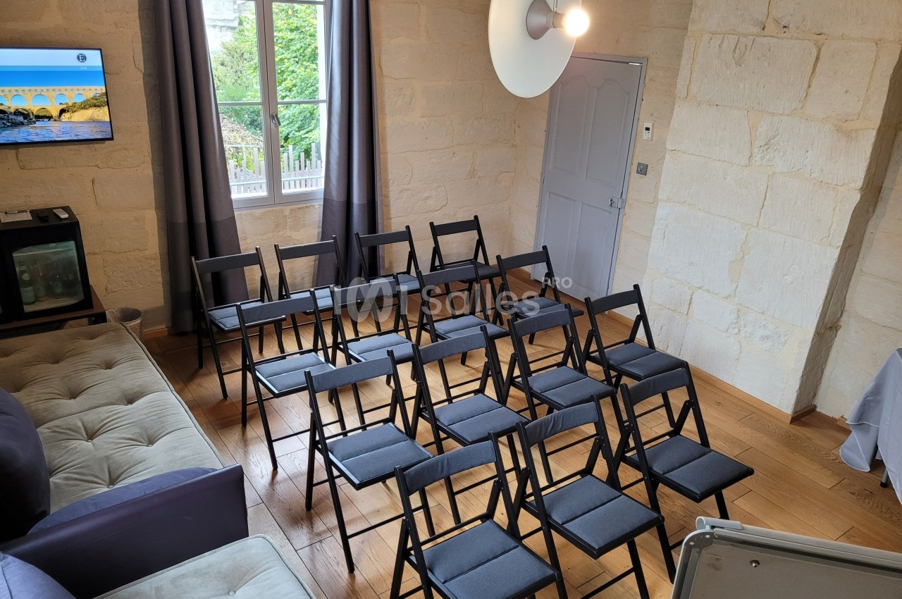 Salle lumineuse avec des chaises pliantes noires disposées en rangées face à une télévision fixée au mur.