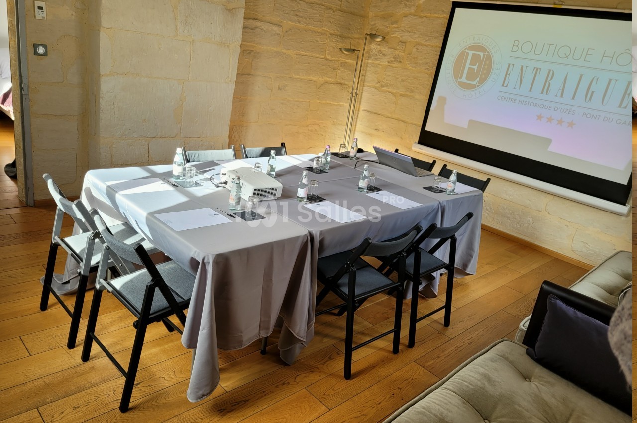 Salle de réunion lumineuse avec table rectangulaire dressée, chaises pliantes, projecteur et écran de présentation.