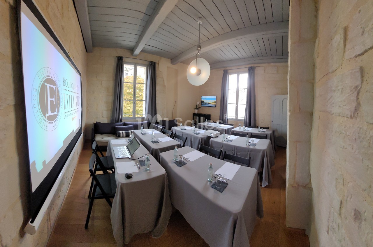 Salle de réunion lumineuse avec tables disposées en rangées, écran de projection allumé et murs en pierre apparente.