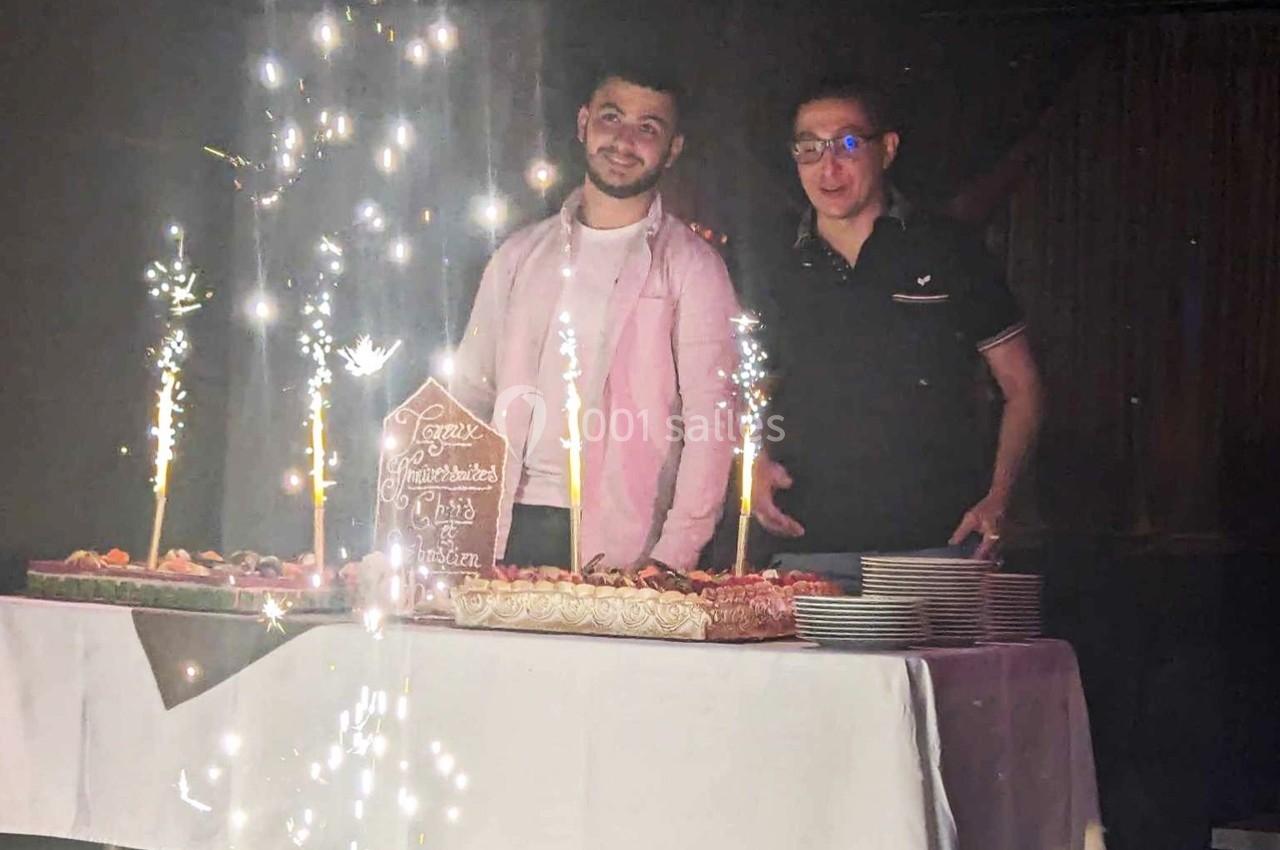 Deux hommes debout derrière une table avec des gâteaux décorés et des cierges étincelants allumés.