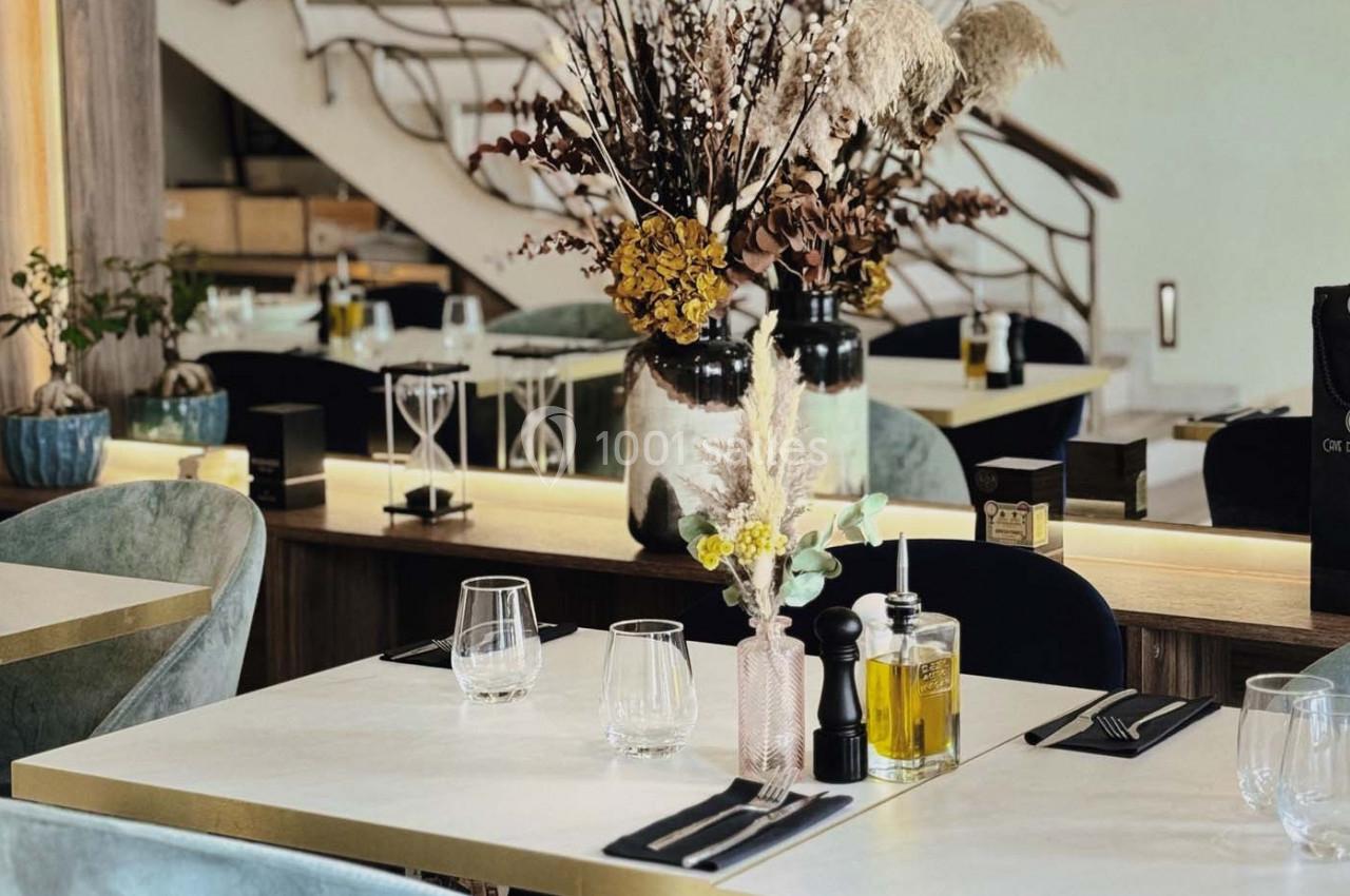 Table dressée avec des verres, couverts et condiments, entourée de chaises, dans un restaurant au décor élégant avec plantes.