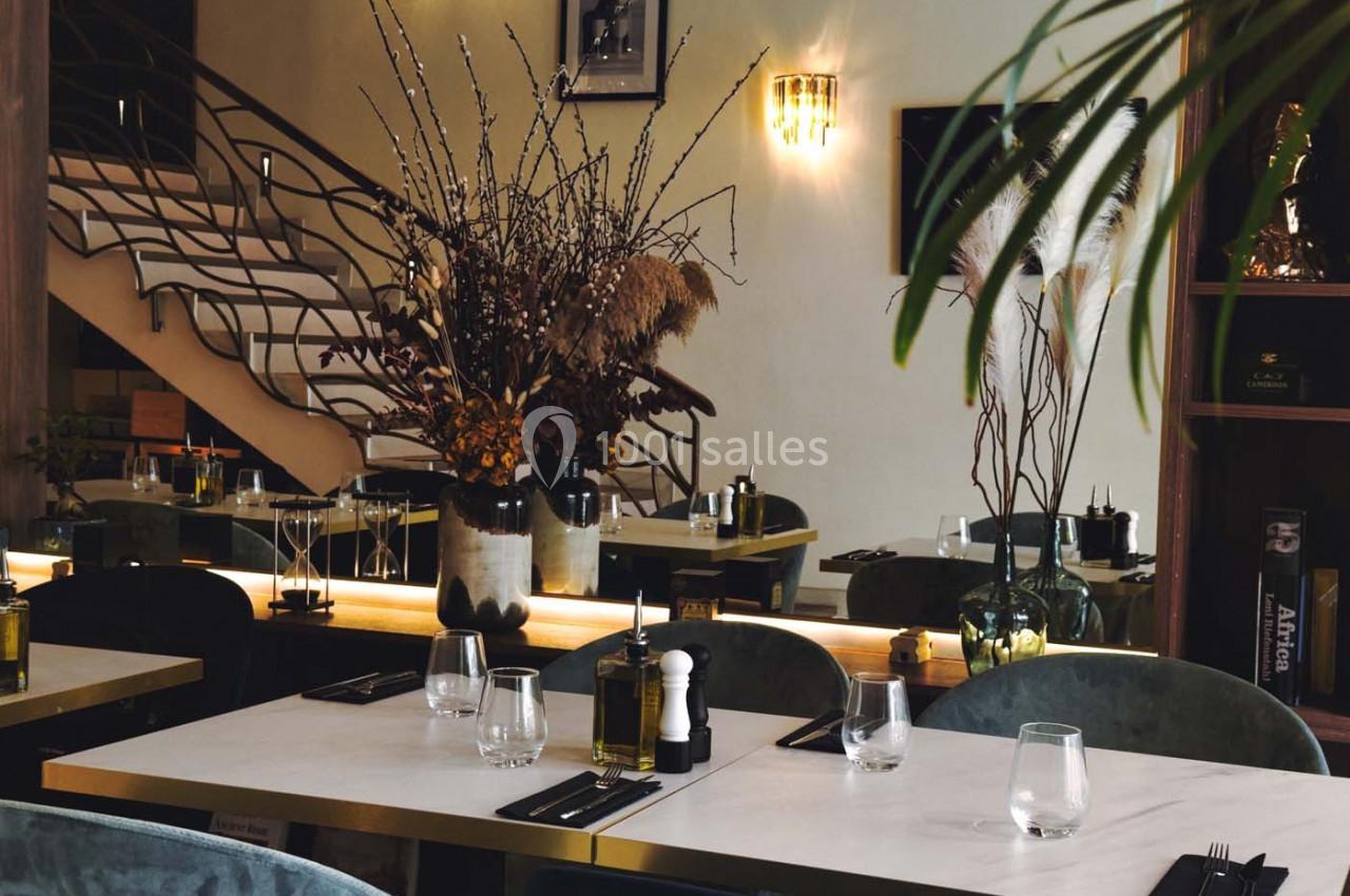 Salle de restaurant élégante avec tables dressées, décorée de plantes et d'un grand bouquet de branches sèches.