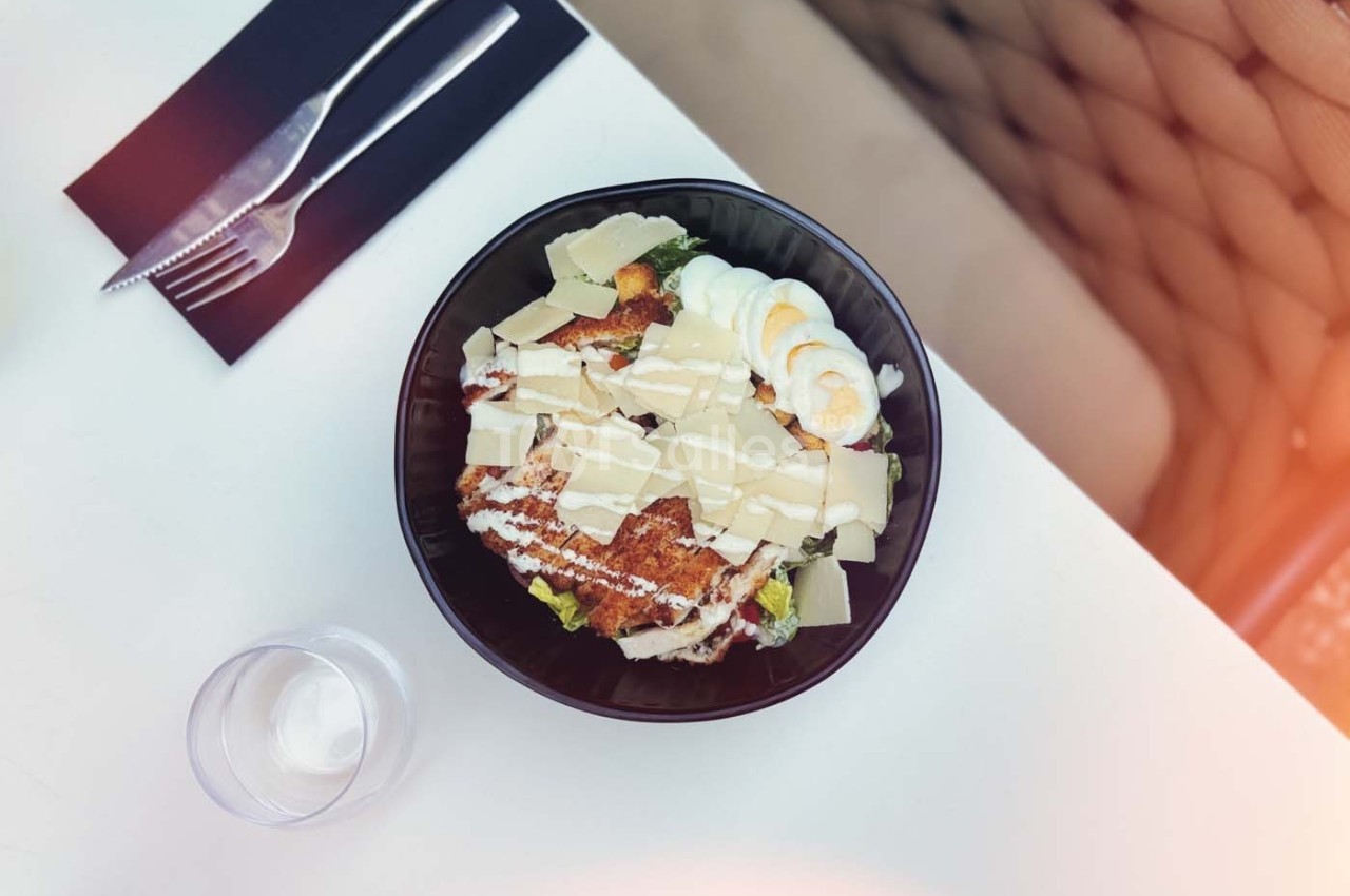 Salade César dans un bol noir avec poulet grillé, œufs durs, copeaux de parmesan et laitue, sur une table blanche.