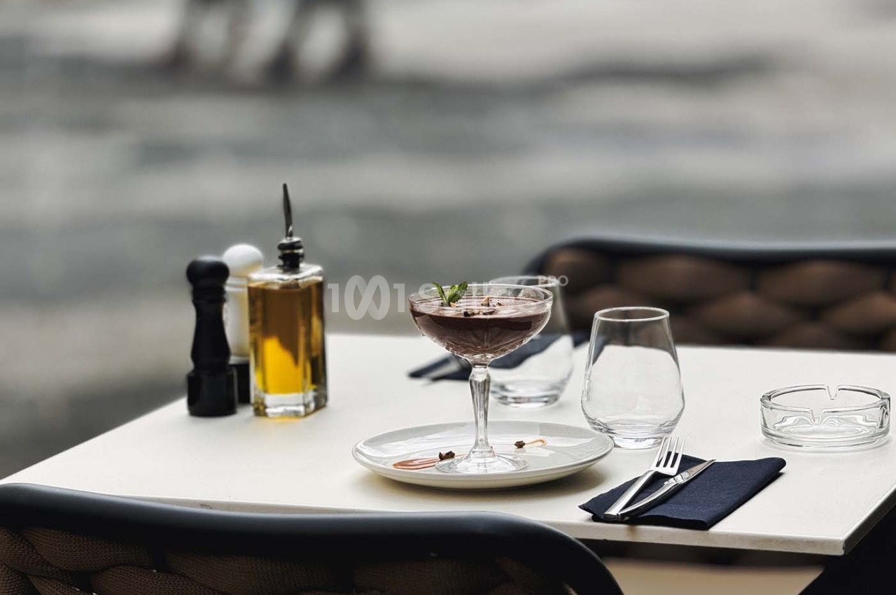 Table de café en extérieur avec un dessert dans une coupe, des verres, une bouteille d'huile et un moulin à poivre.