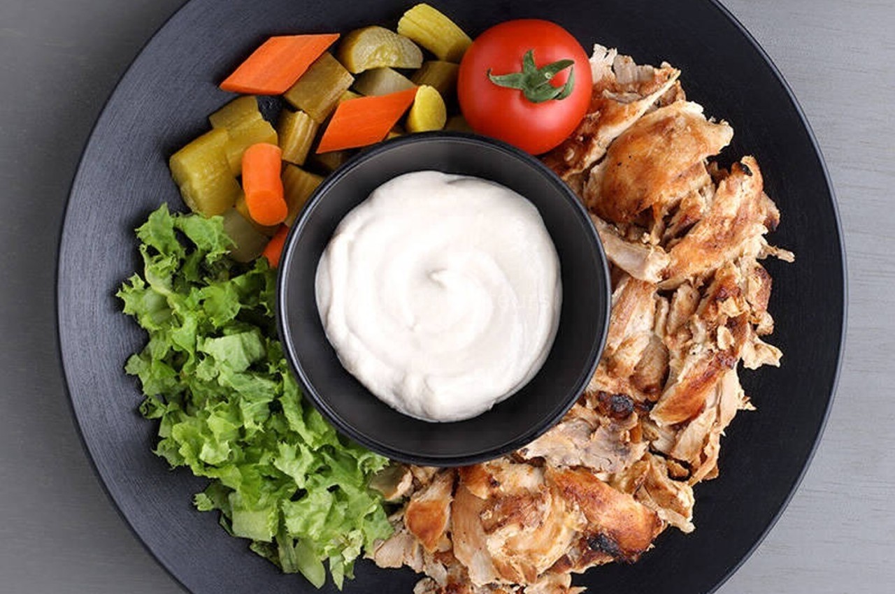 Assiette contenant du poulet grillé, sauce blanche, salade verte, légumes marinés et une tomate cerise.