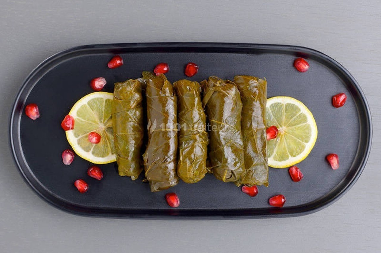 Feuilles de vigne farcies alignées sur une assiette noire avec rondelles de citron et graines de grenade.
