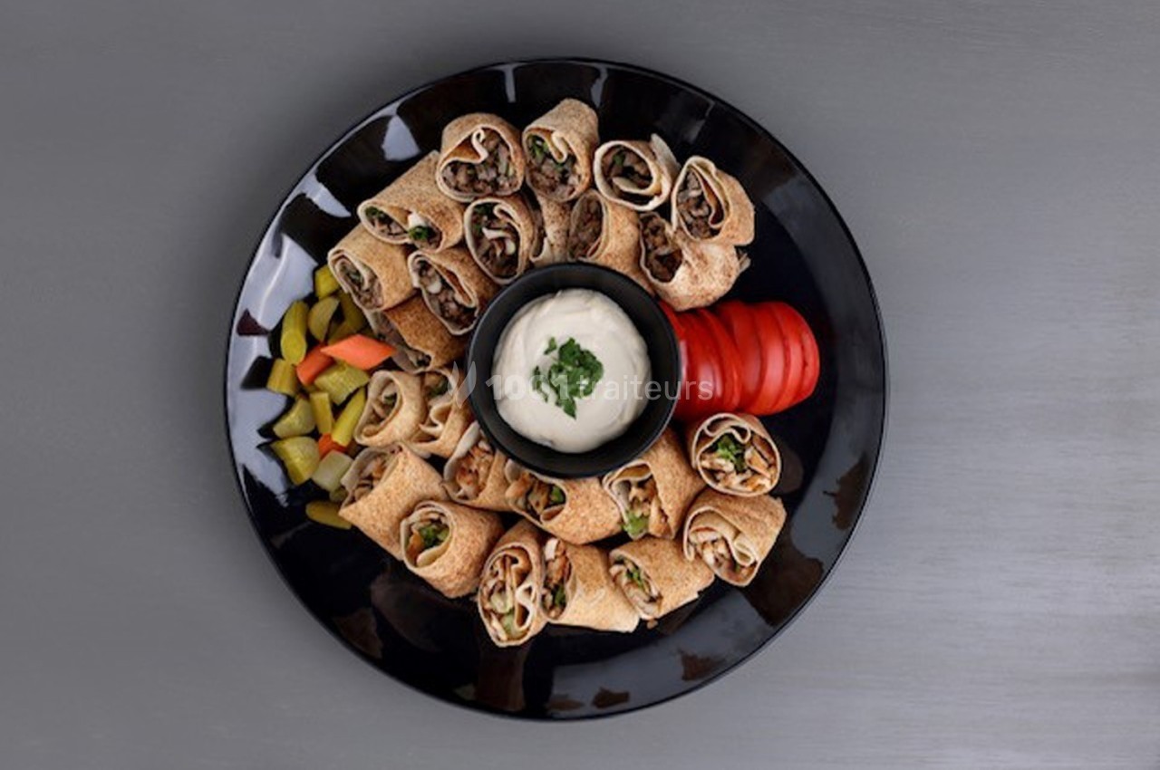 Assiette noire contenant des wraps garnis, accompagnée de légumes, de tranches de tomate et d'une sauce blanche au centre.