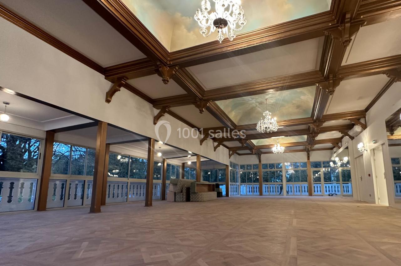 Salle spacieuse avec parquet en bois, plafond à caissons décoré de fresques et lustres, entourée de grandes fenêtres.