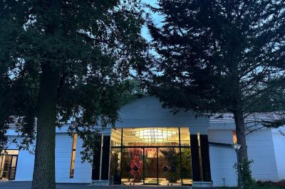 Entrée d'un bâtiment blanc entouré d'arbres, avec des portes vitrées illuminées par une lumière intérieure.