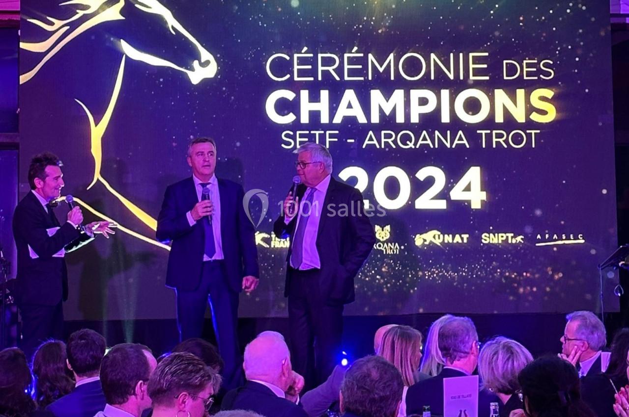 Deux hommes sur scène s'expriment lors d'une cérémonie avec un écran affichant ’Cérémonie des Champions 2024’.