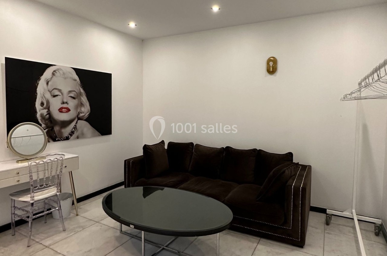 Salon minimaliste avec canapé marron, table basse en verre, coiffeuse blanche, portant à vêtements et portrait mural.