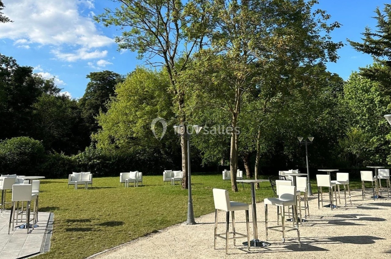 Tables hautes et chaises blanches disposées sur une terrasse et une pelouse, entourées d'arbres par temps ensoleillé.