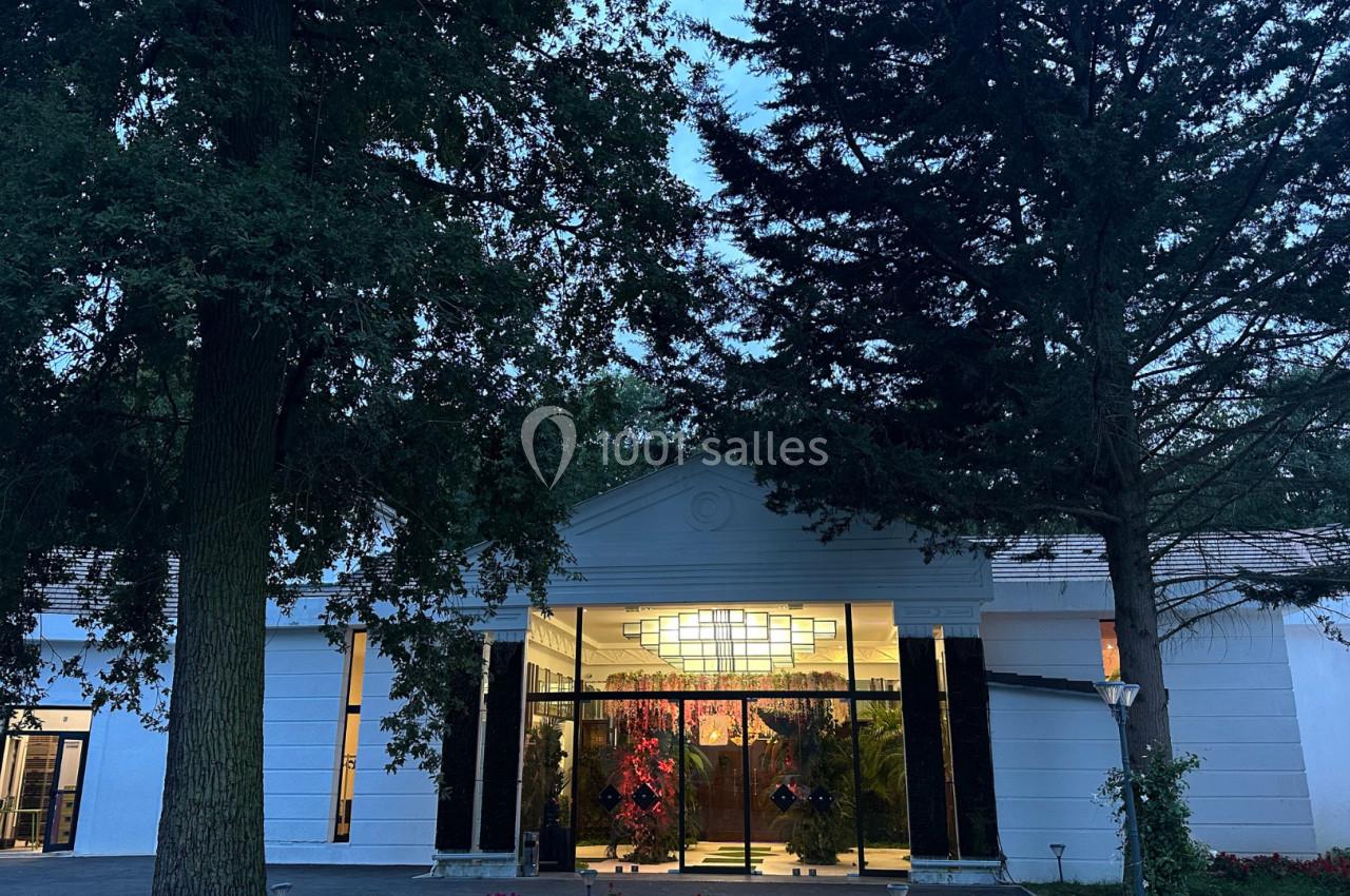 Entrée d'un bâtiment blanc entouré d'arbres, avec des portes vitrées éclairées et un lustre visible à l'intérieur.