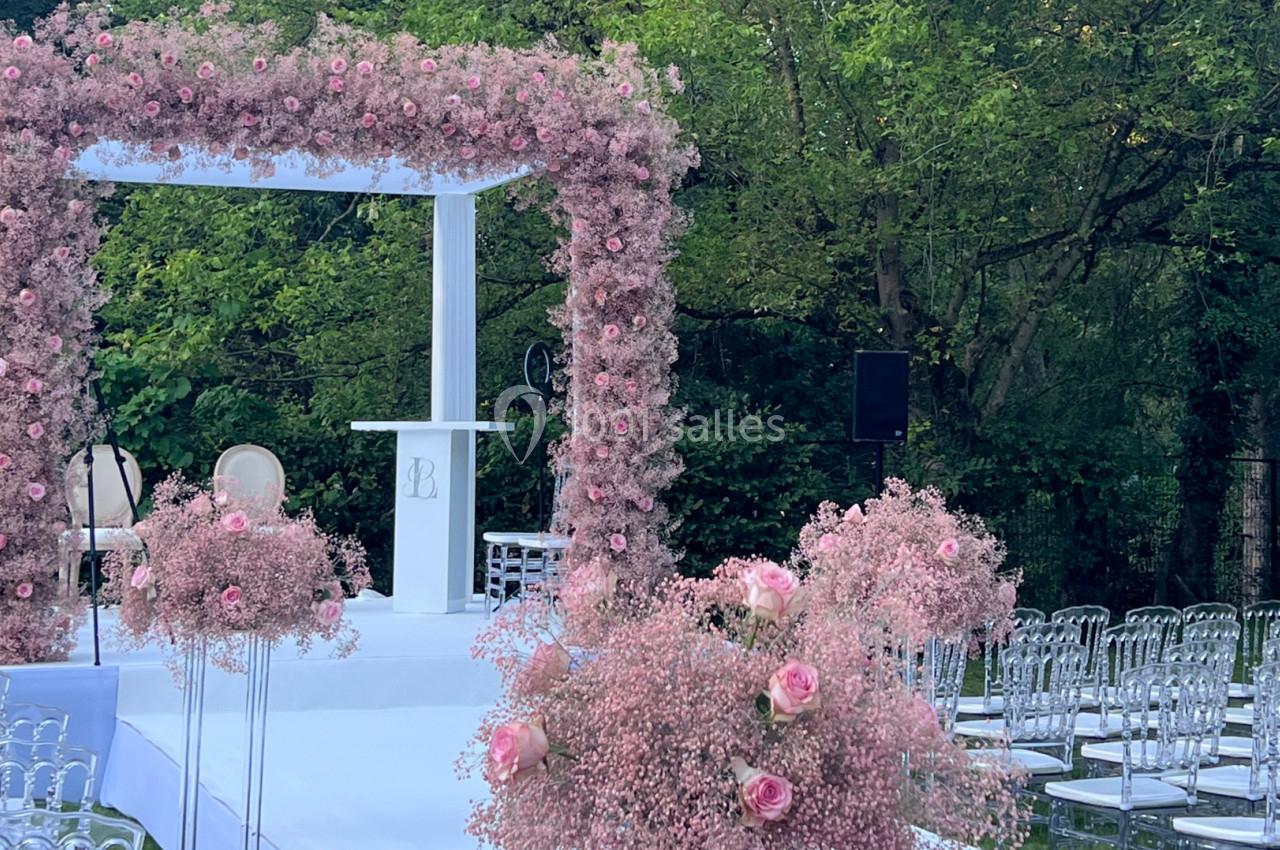 Arche de cérémonie ornée de fleurs roses, entourée de chaises transparentes, dans un jardin verdoyant.