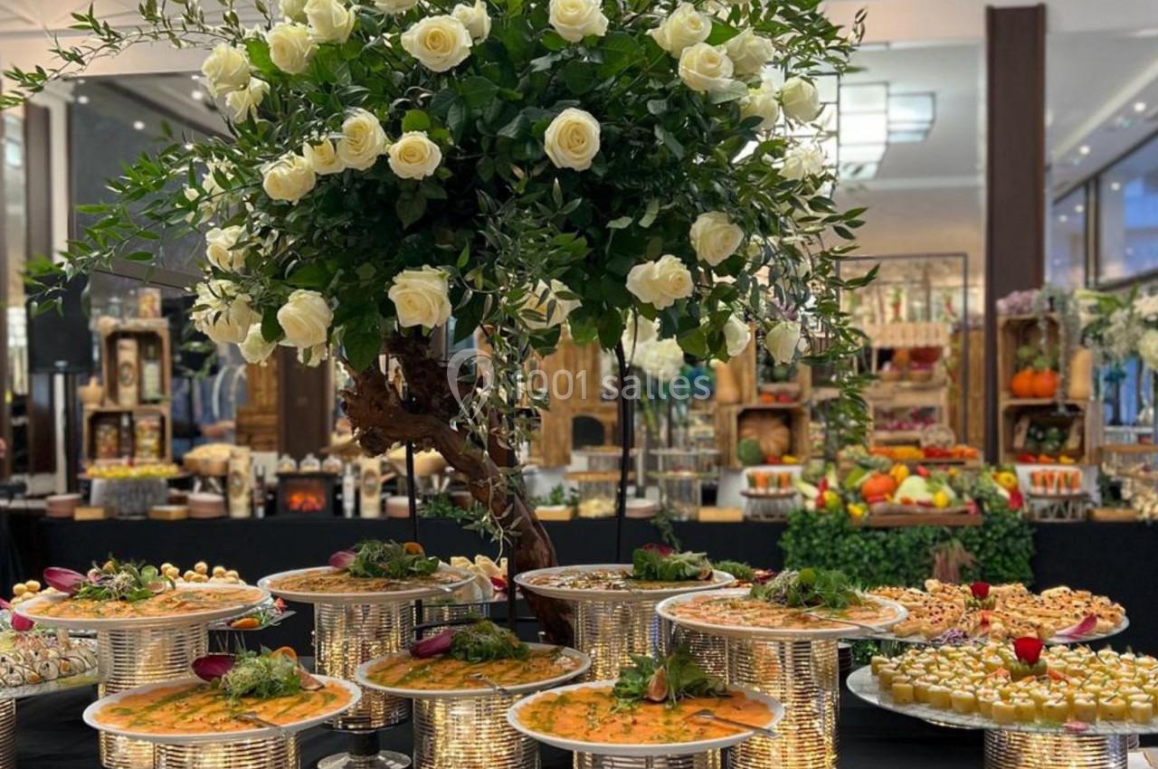 Buffet élégant avec plats variés sur des présentoirs argentés, décoré d'un grand bouquet de roses blanches.