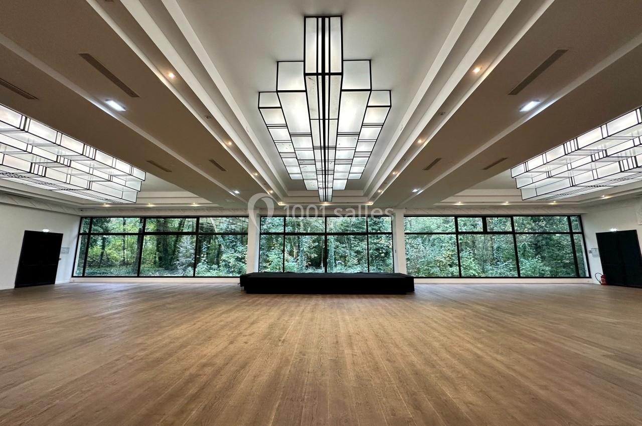 Salle spacieuse avec parquet clair, grandes baies vitrées donnant sur des arbres et plafonniers géométriques modernes.