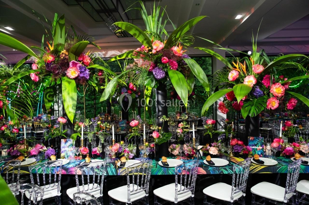 Table dressée avec des chaises transparentes, ornée de compositions florales tropicales colorées et de bougies.