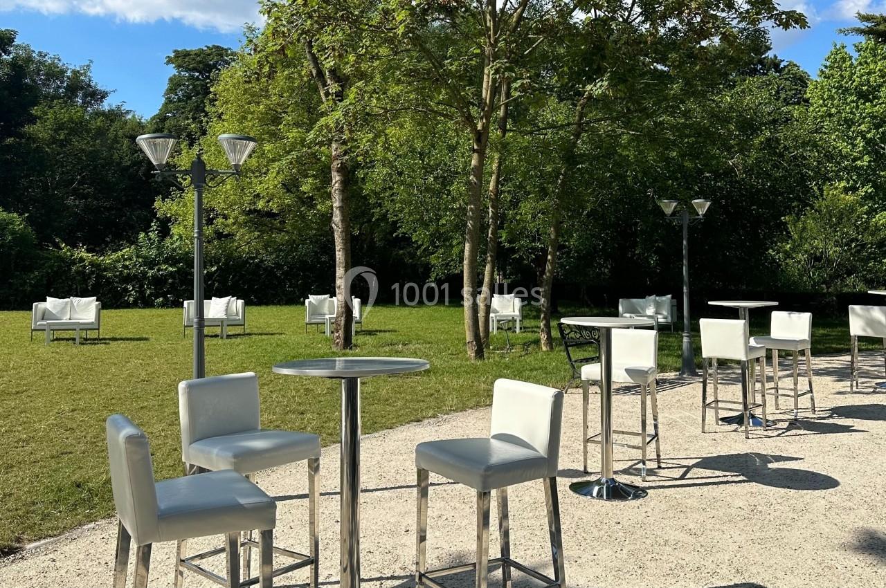 Chaises et tables hautes blanches disposées sur une allée en gravier, entourées de pelouse et d'arbres.