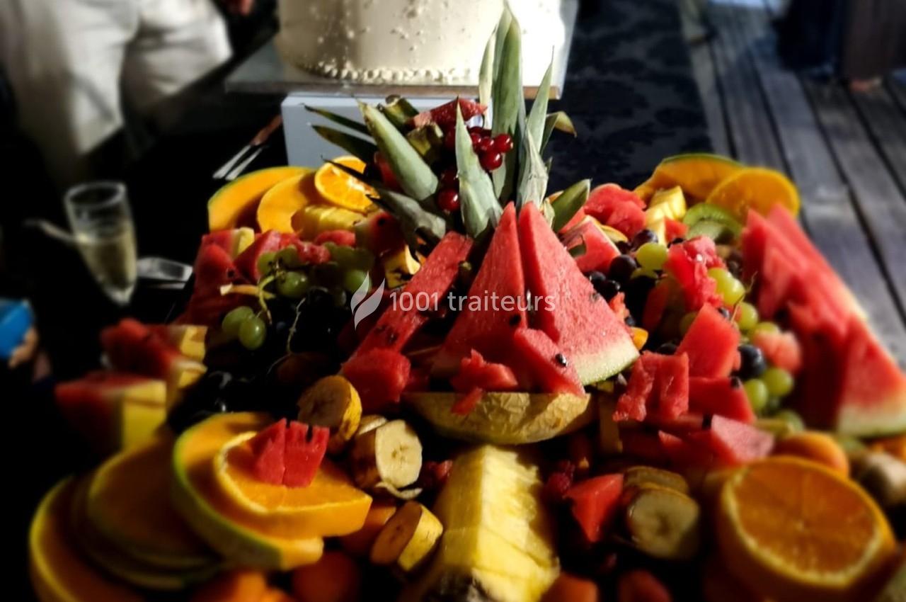 Plateau de fruits variés avec pastèques, oranges, bananes, raisins et ananas, disposé devant un gâteau blanc.