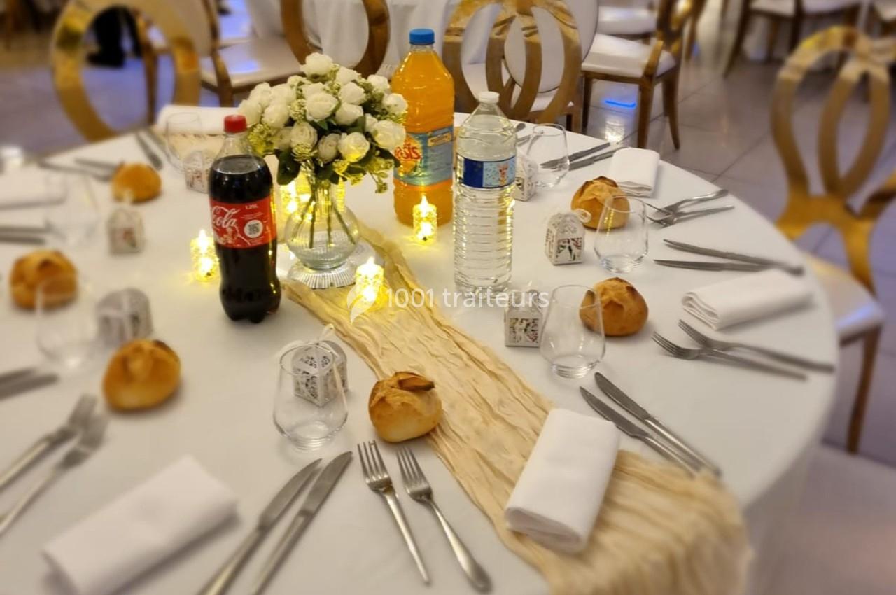 Table dressée pour un repas avec nappes blanches, couverts, pains, bouteilles de boissons et décoration florale centrale.