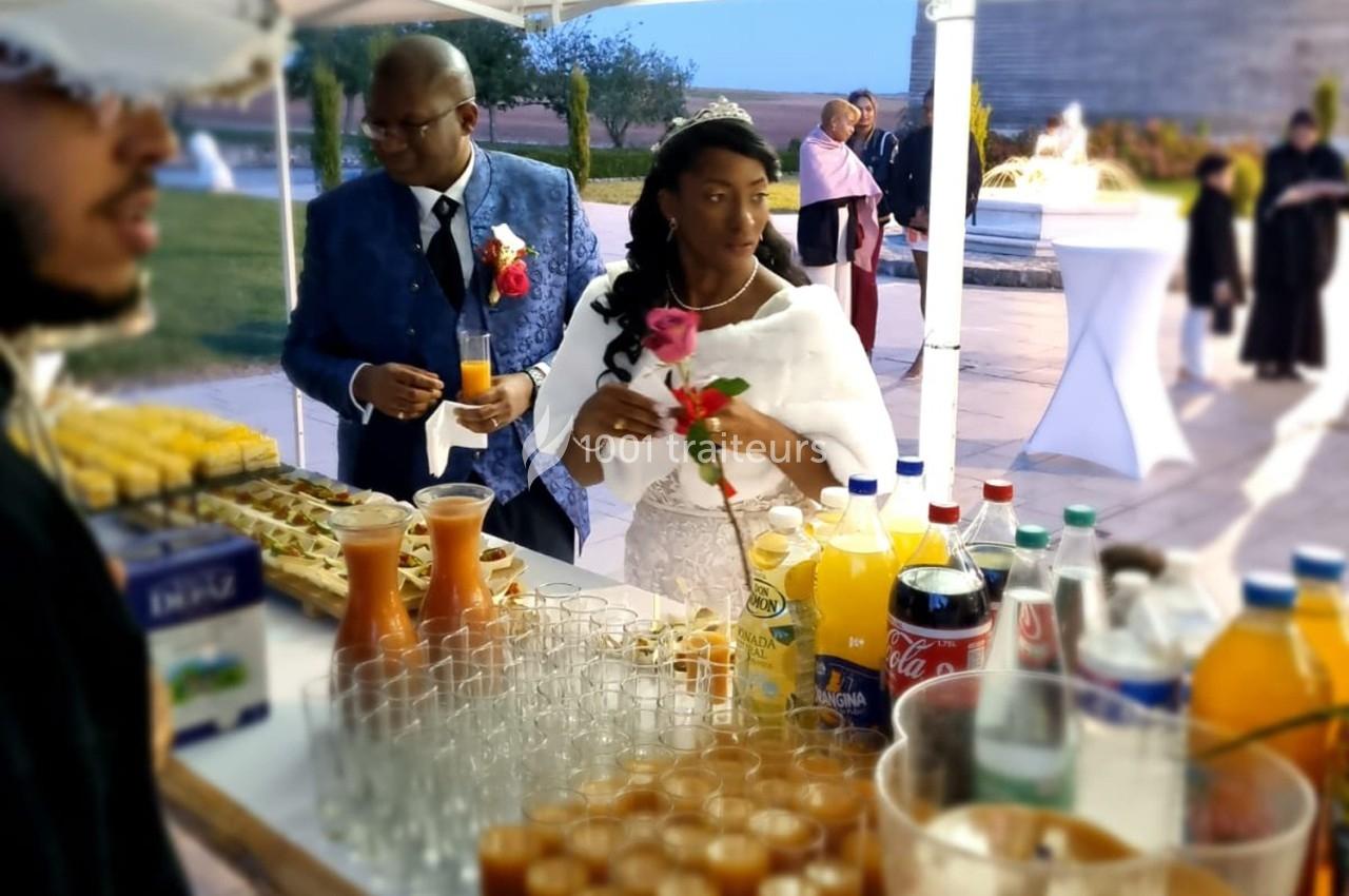 Un couple en tenue de mariage se sert à un buffet en plein air, entouré de boissons et de verrines.
