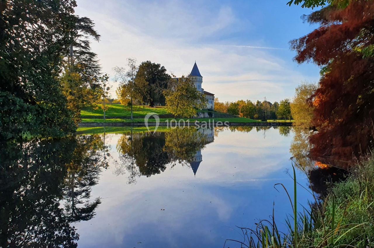 Vue d'un château entouré d'arbres, reflété dans un étang calme sous un ciel dégagé.