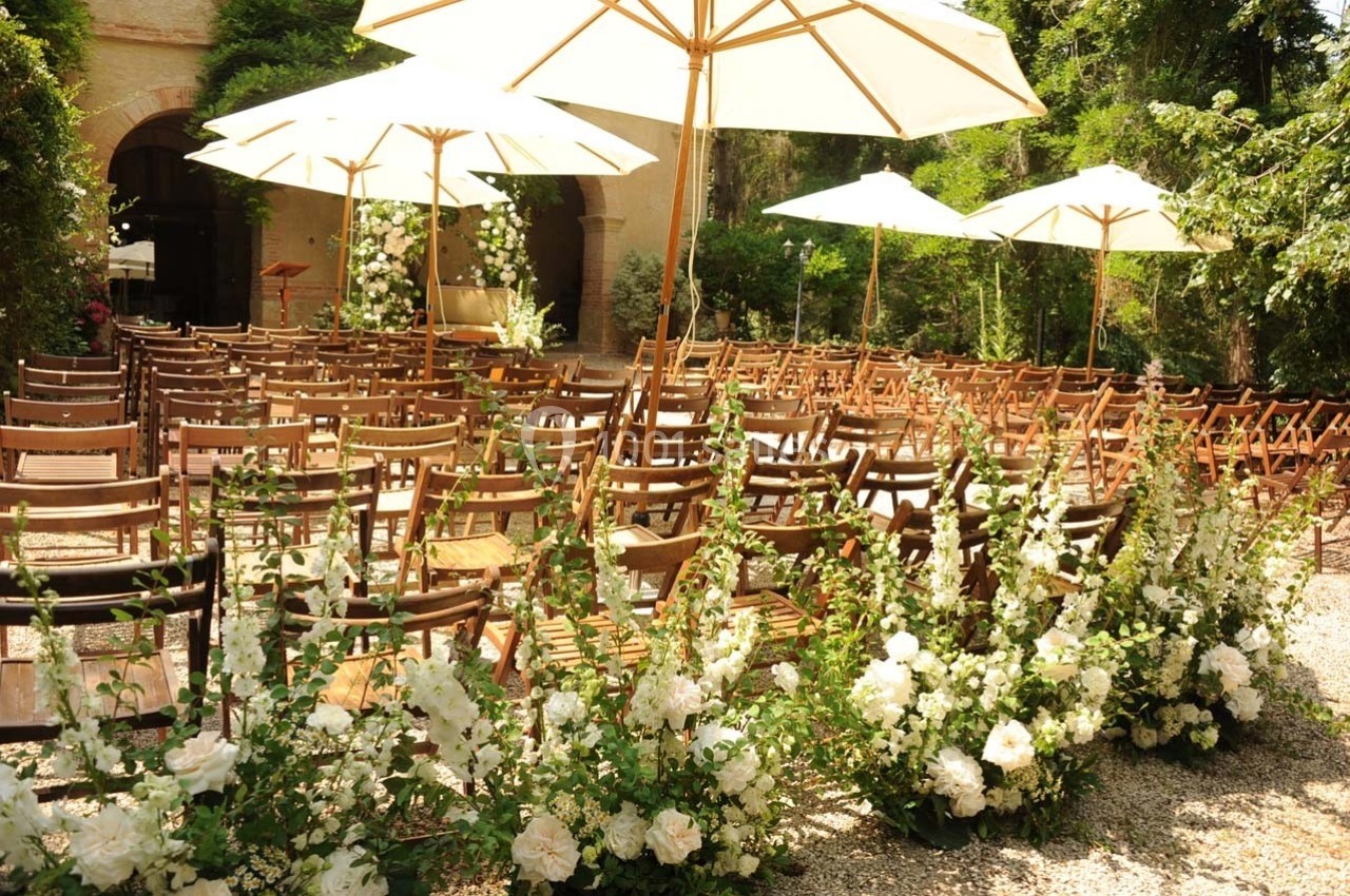 Chaises en bois alignées sous des parasols blancs, entourées de fleurs blanches, dans un jardin ensoleillé.