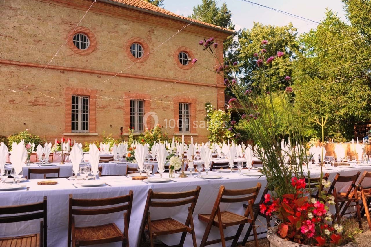 Grande table extérieure dressée avec nappes blanches et vaisselle, devant un bâtiment en briques et un jardin fleuri.