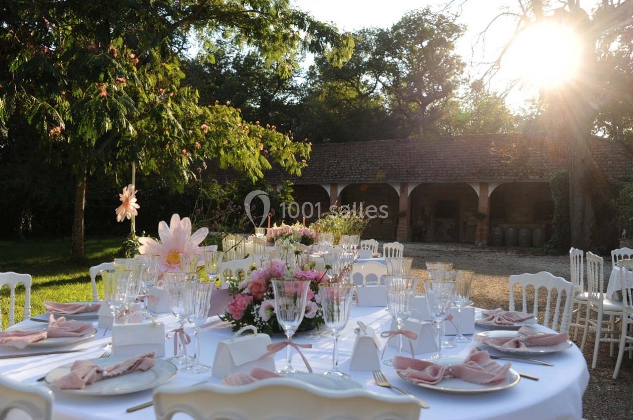 Table dressée en extérieur avec nappes blanches, vaisselle élégante et décorations florales, sous un soleil couchant.
