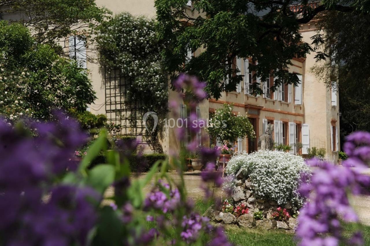 Façade d'une maison entourée de végétation, avec des fleurs violettes et blanches au premier plan.