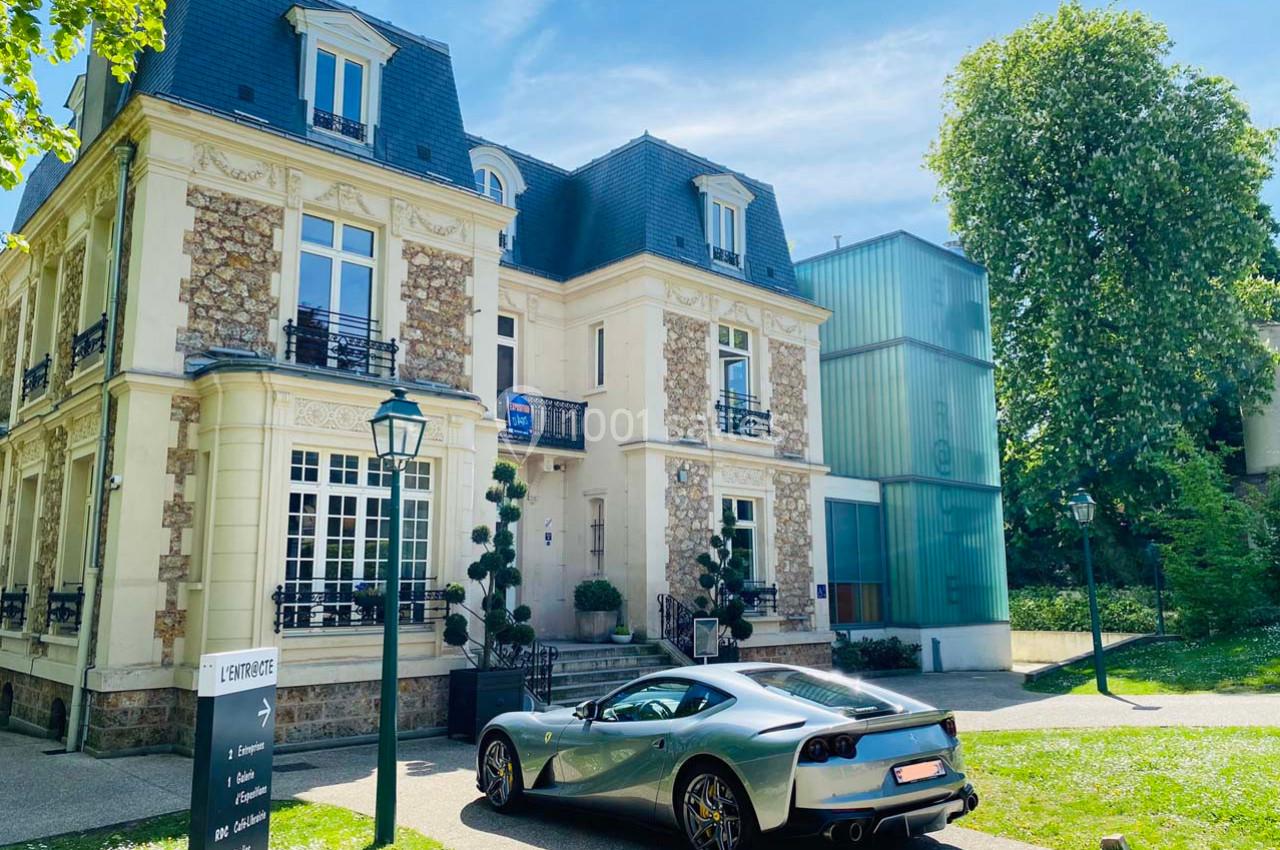 Voiture de sport grise stationnée devant une maison bourgeoise en pierre et un bâtiment moderne en verre.