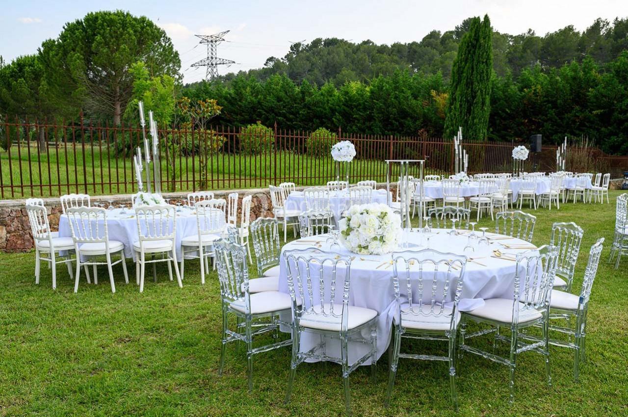 Tables rondes décorées de nappes blanches et de fleurs, disposées dans un jardin pour un événement en plein air.
