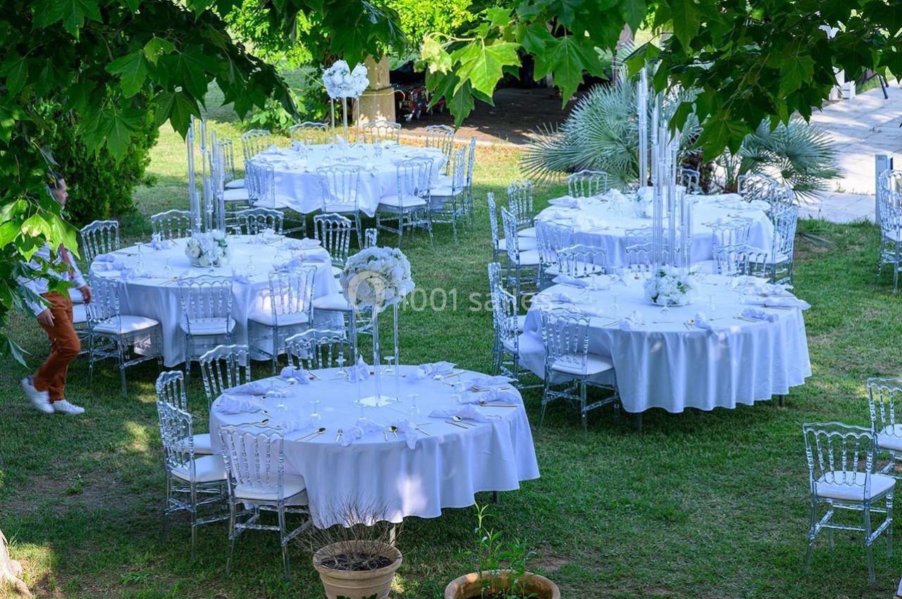 Tables rondes dressées avec nappes blanches et décorations florales, disposées dans un jardin verdoyant.