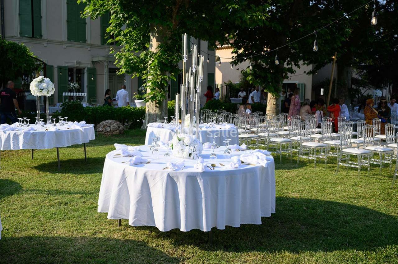 Tables rondes dressées avec nappes blanches dans un jardin, entourées de chaises transparentes et décorées de bougies.