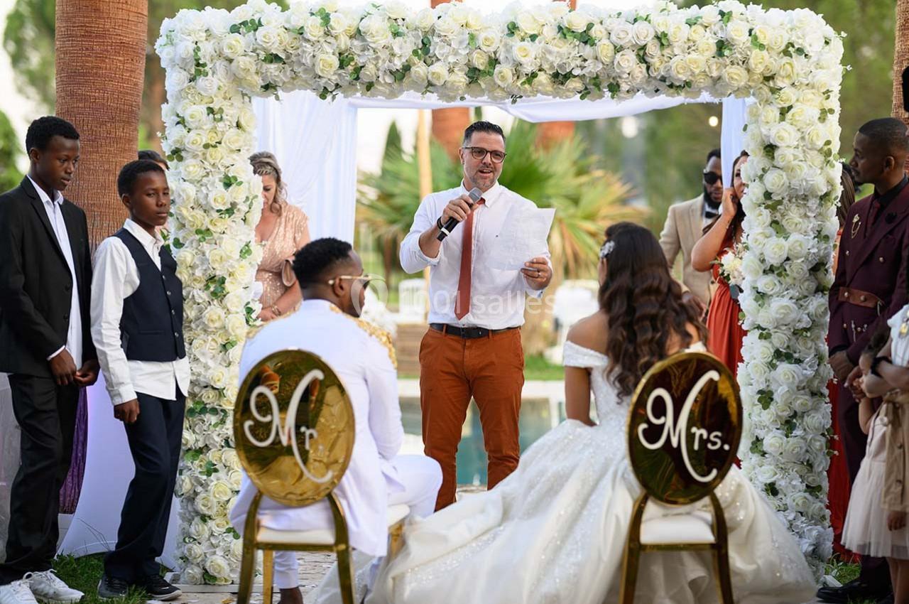 Un couple assis face à un officiant lisant un discours lors d'une cérémonie de mariage en extérieur, entouré d'invités.