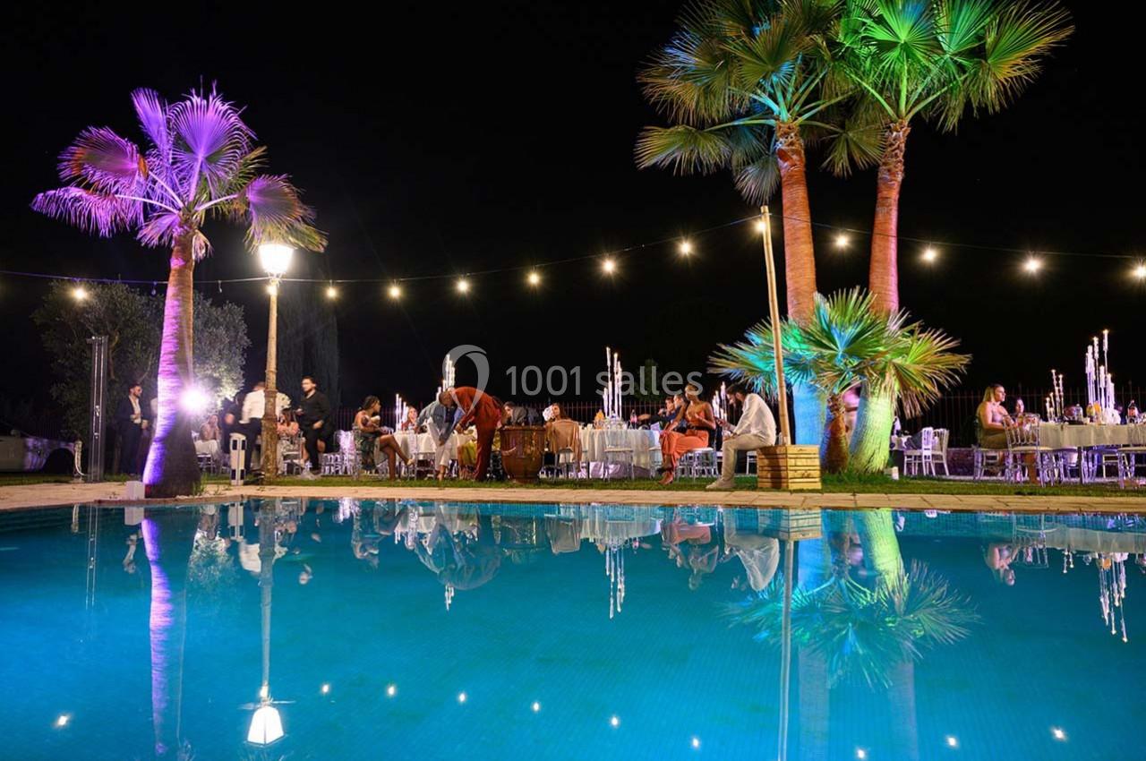 Vue nocturne d'une réception en plein air avec des invités assis près d'une piscine éclairée et entourée de palmiers.