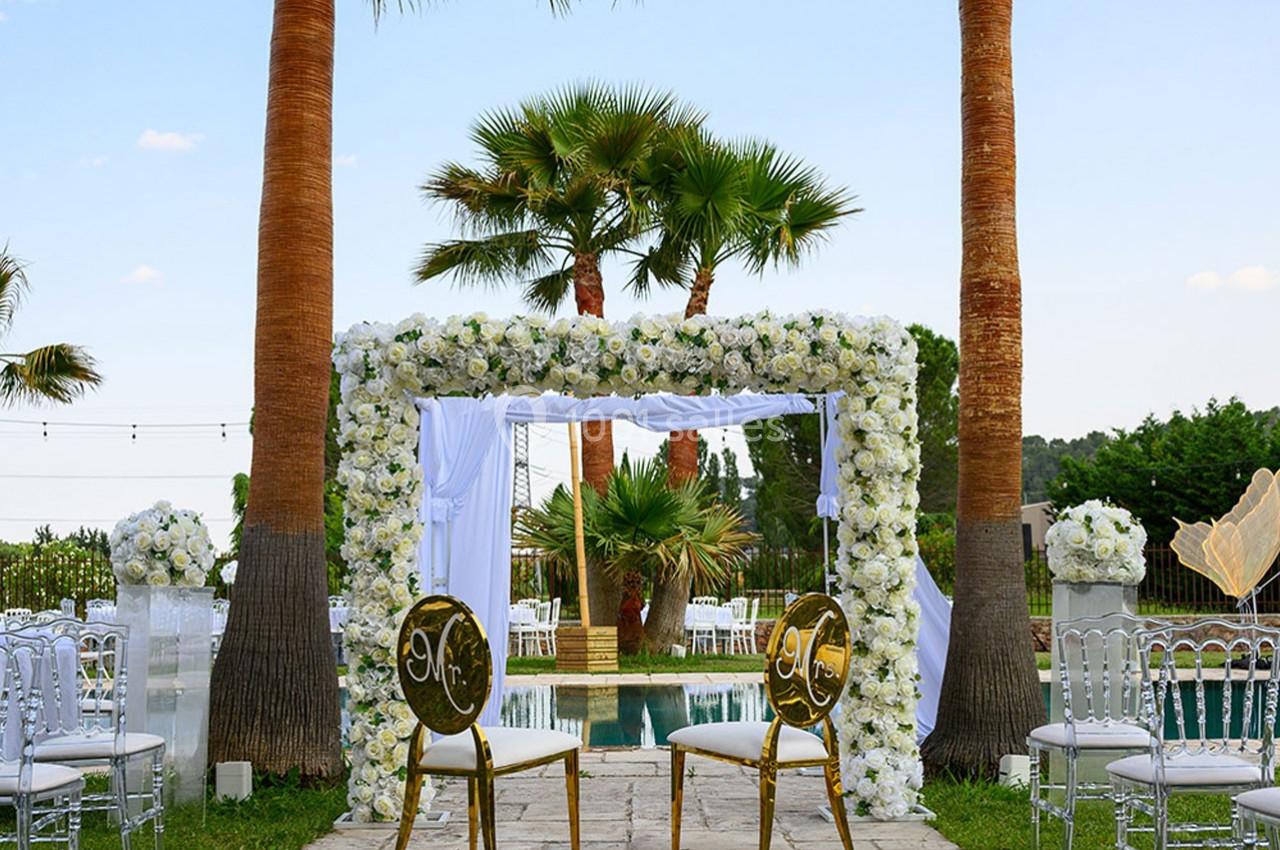 Arche de mariage décorée de fleurs blanches, entourée de palmiers, avec deux chaises dorées devant une piscine.