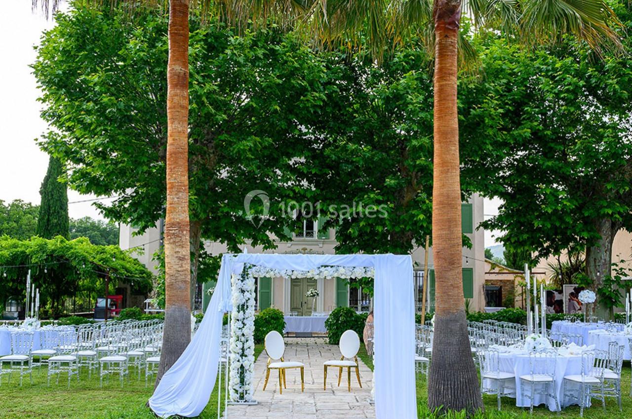 Décoration de mariage en extérieur avec arche blanche, chaises et tables dressées sous des arbres et des palmiers.