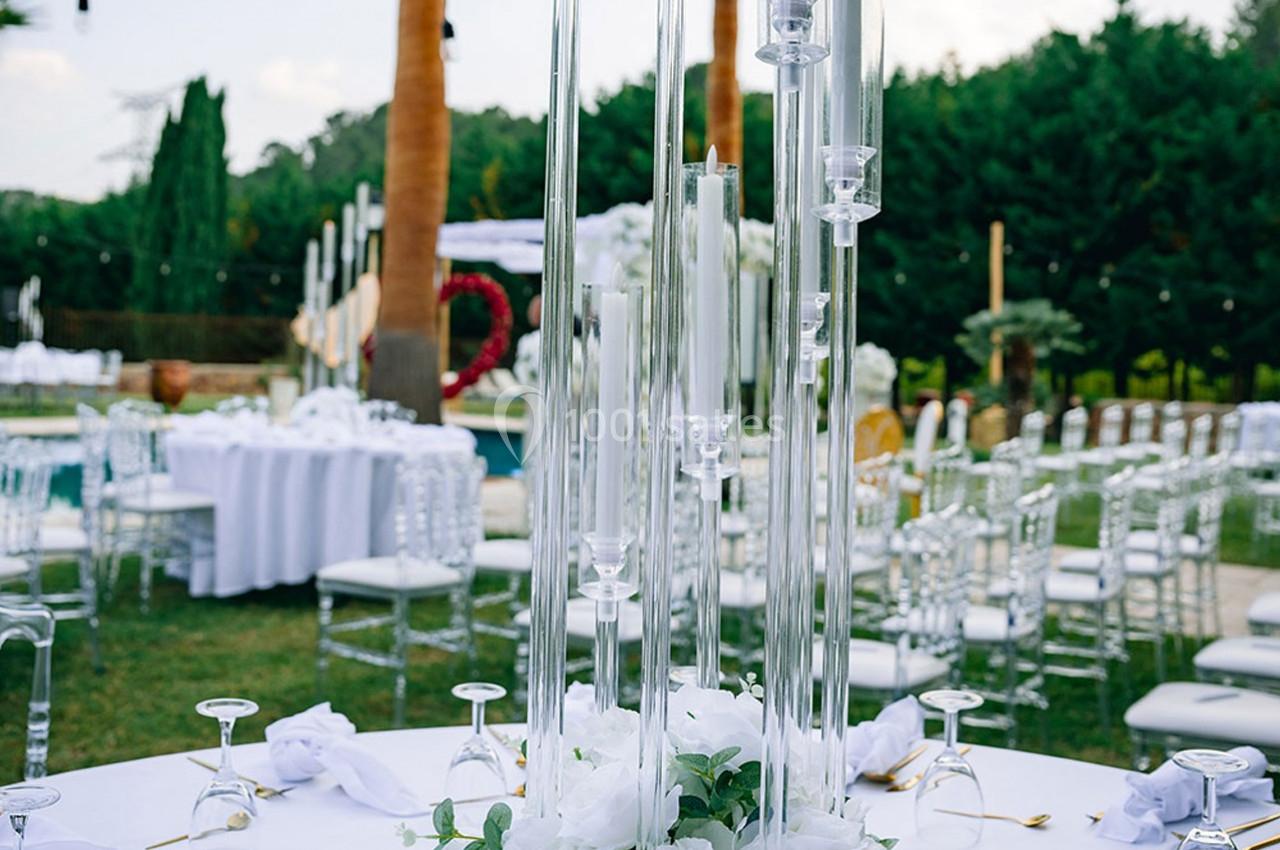 Décoration de table de mariage en extérieur avec chandeliers en verre, fleurs blanches et chaises alignées sur une pelouse.
