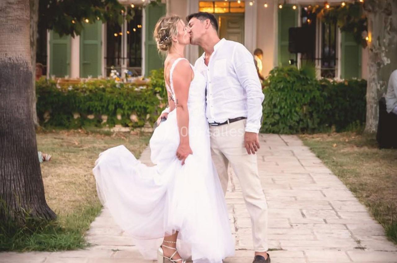 Un couple en tenue de mariage s'embrasse dans une allée pavée, entourée de verdure et de lumières suspendues.