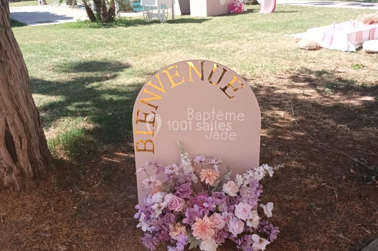 Panneau ’Bienvenue Baptême Jade’ décoré de fleurs roses et violettes, installé dans un jardin ensoleillé.
