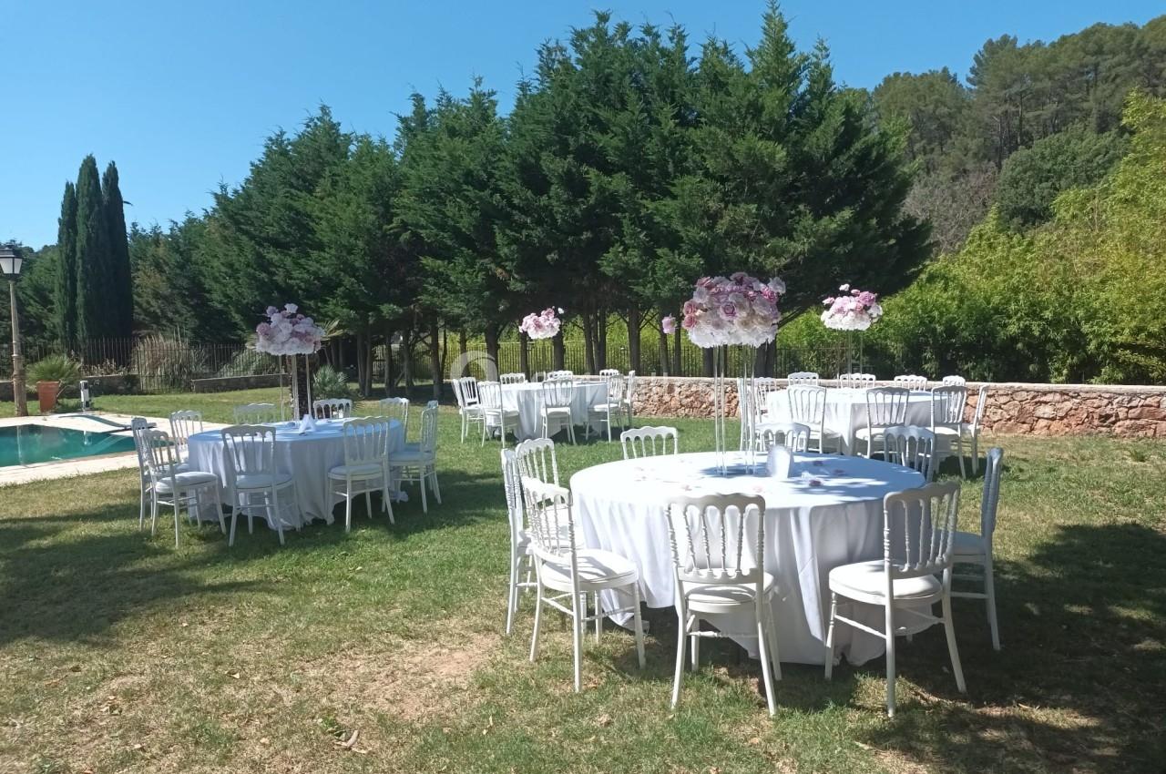 Tables rondes décorées de nappes blanches et centres de table fleuris, disposées dans un jardin verdoyant.
