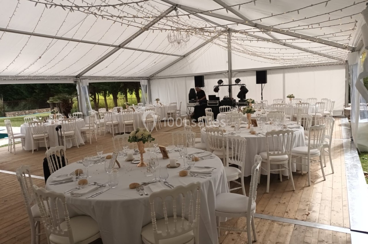 Salle de réception sous chapiteau avec tables rondes dressées, chaises blanches et guirlandes lumineuses suspendues.