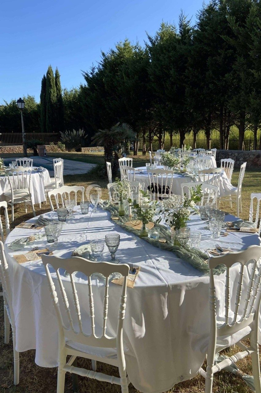 Tables rondes dressées avec nappes blanches, vaisselle et décor floral, disposées en extérieur près d'arbres et d'une…