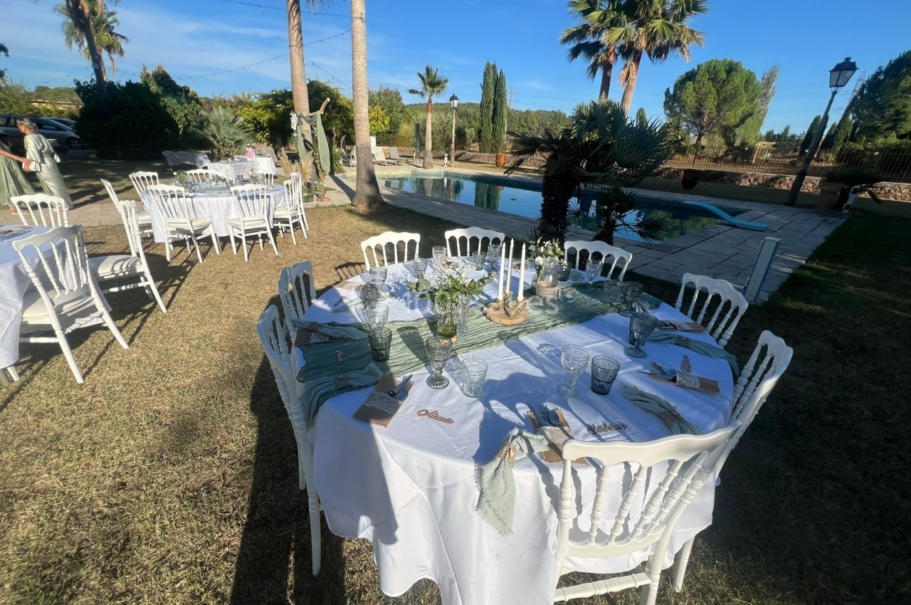 Table dressée en extérieur avec nappes blanches, décorations florales et vue sur un jardin avec piscine.