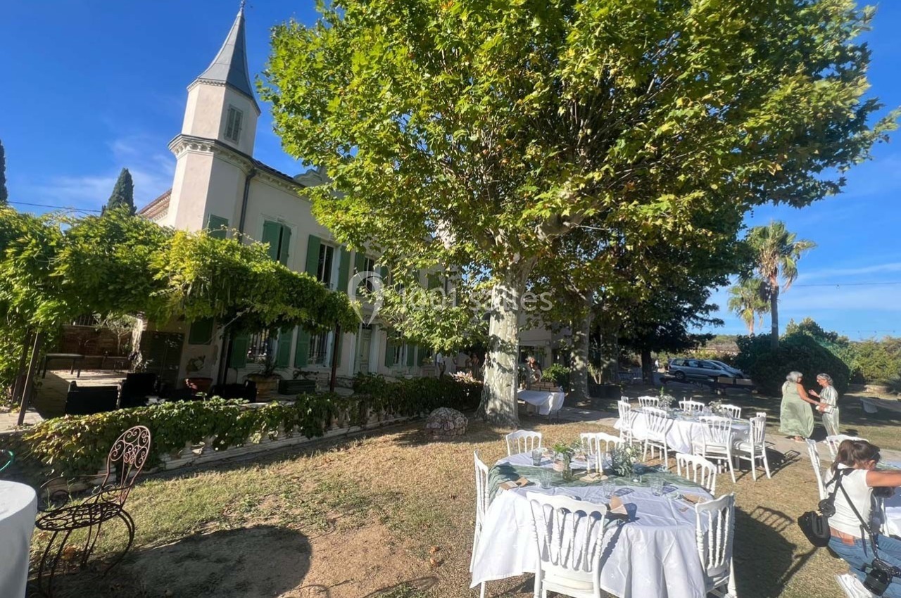 Tables dressées dans un jardin ombragé devant une grande maison avec des volets verts et une petite tour.
