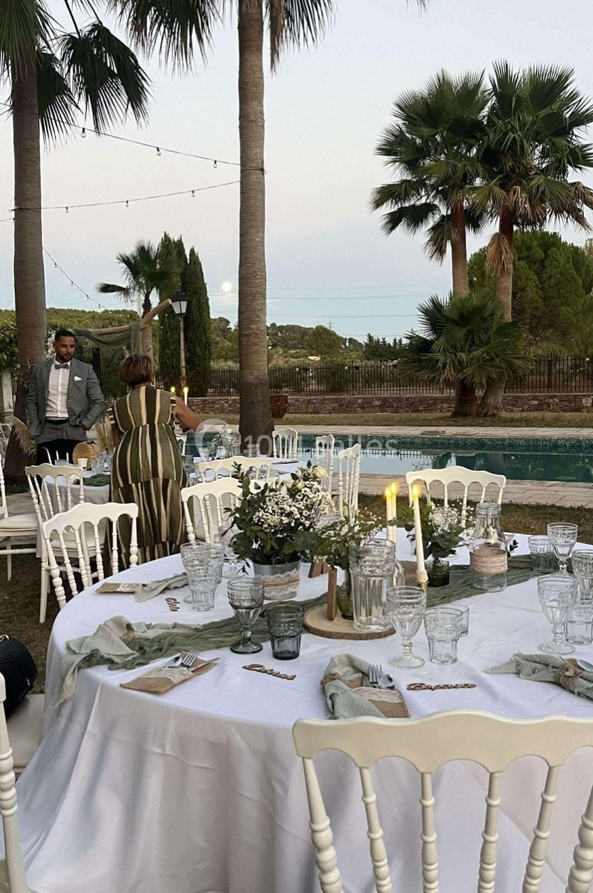 Tables dressées en extérieur avec nappes blanches, décorations florales, bougies et palmiers en arrière-plan.