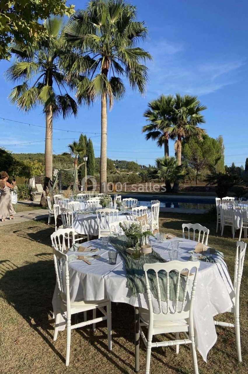 Tables dressées en extérieur avec nappes blanches, entourées de chaises, près d'une piscine et de palmiers sous un ciel bleu.